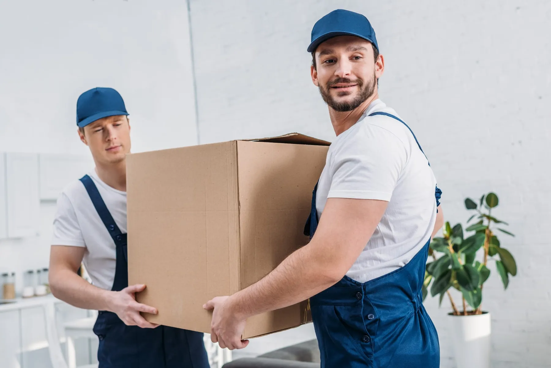 Two movers in blue caps and overalls carrying a large cardboard box